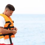 Handsome male lifeguard putting on life vest near sea