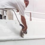 Yacht maintenance. A man polishing side of the white boat by grinder machine in the marina